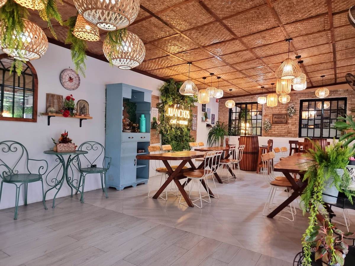 An inviting dining area features a mix of wooden and metal furniture, allowing for flexible seating arrangements. Soft lighting is provided by woven pendant lamps, and greenery adds a refreshing touch. Decorative elements, including a vintage clock and artfully arranged shelves, enhance the ambiance.