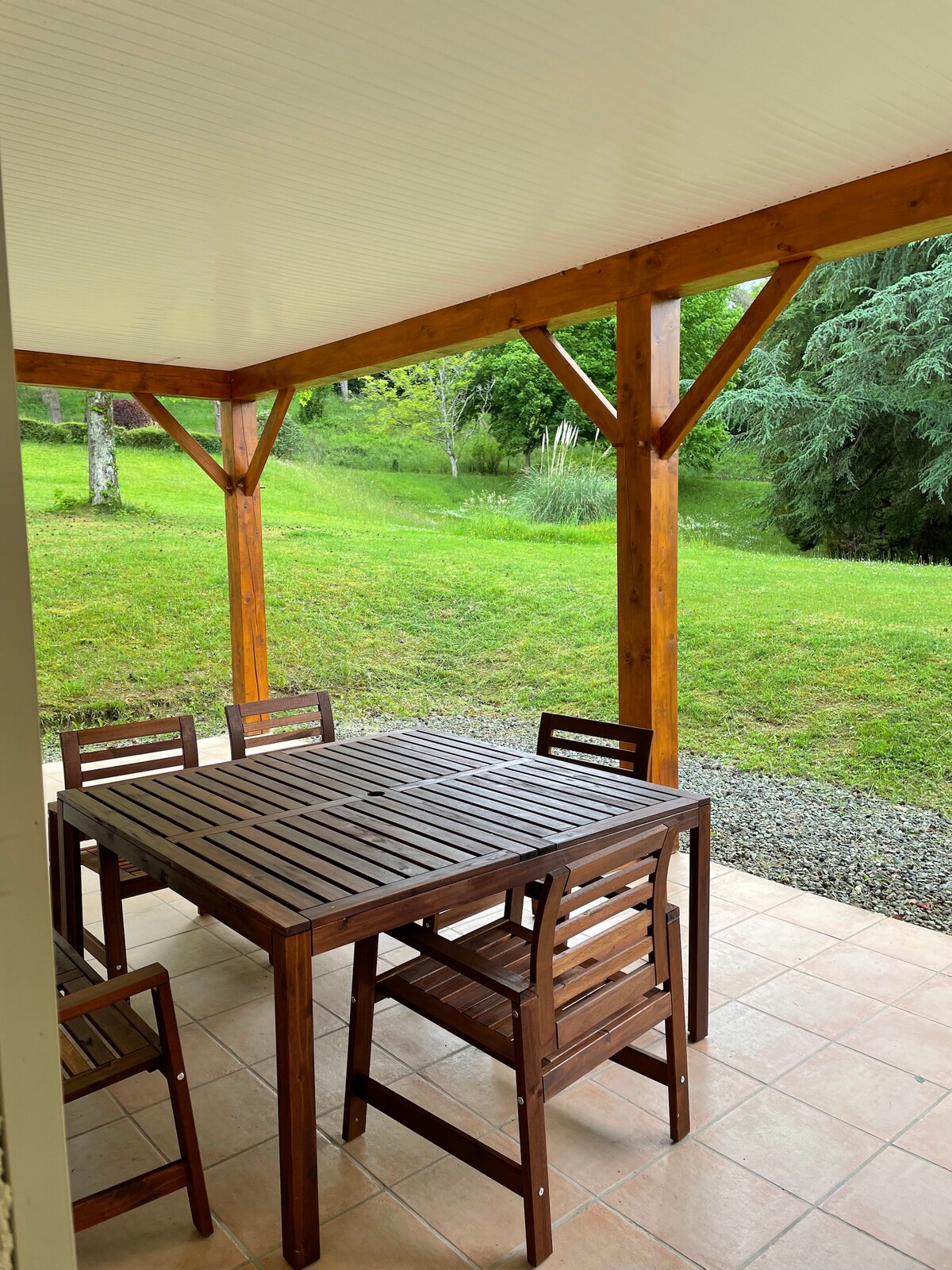An outdoor dining area is featured under a wooden pergola, showcasing a large table surrounded by six wooden chairs. The surrounding green garden is visible, enhancing the natural setting, with grass and trees in the background.