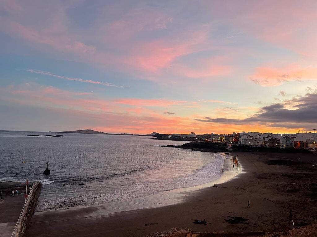 A serene beach scene at sunset is captured, featuring gentle waves lapping the shore. The sky displays a gradient of soft pinks and blues, while distant hills and buildings provide a subtle backdrop. A peaceful ambiance is created, inviting moments of relaxation and reflection.