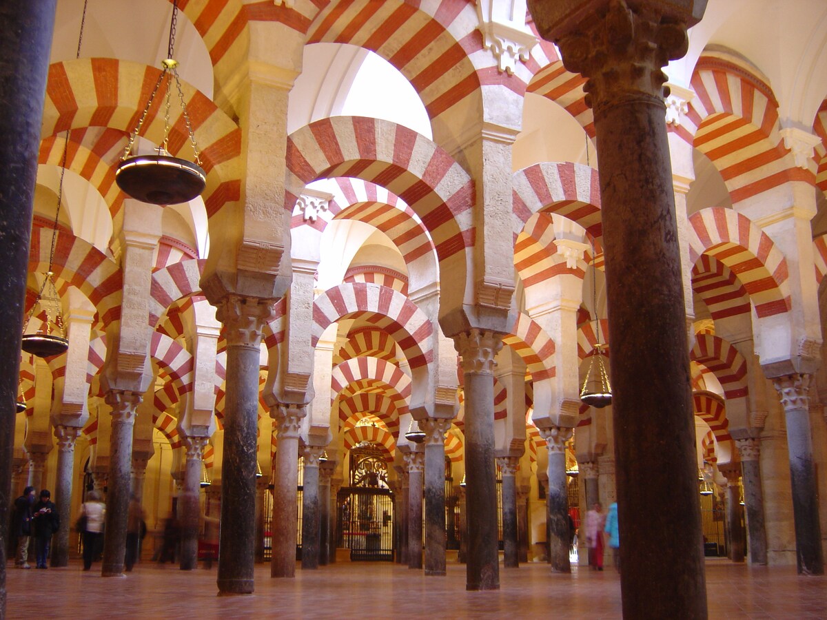 An expansive hall showcases a series of columns adorned with intricate patterns, supporting arches of alternating red and white stripes. The well-lit space features a warm wooden floor, with glimpses of visitors moving through, highlighting the grandeur of the architectural design.