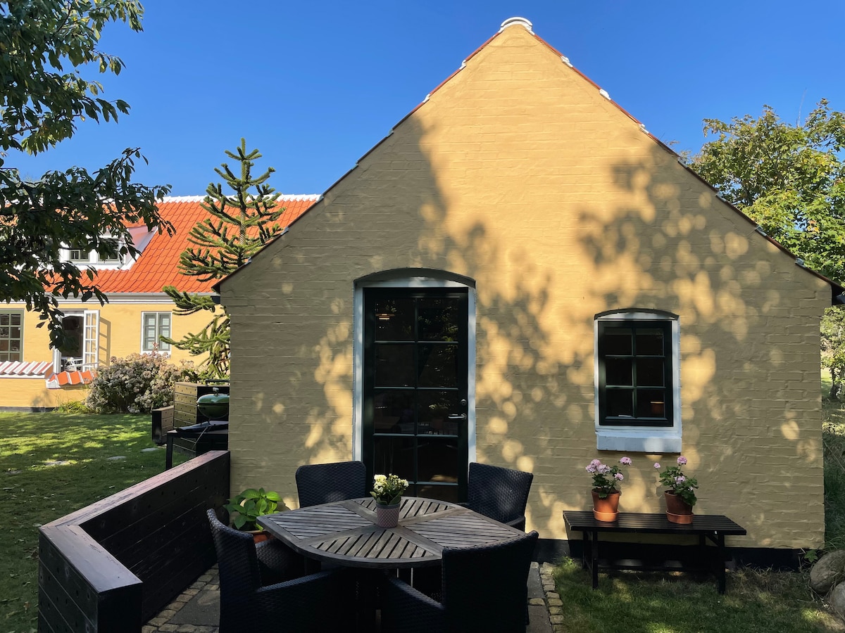 A charming yellow exterior of the annex is showcased, featuring large windows that invite natural light. A circular wooden dining table surrounded by black wicker chairs is positioned in the foreground, and potted plants add a touch of greenery on a nearby bench.