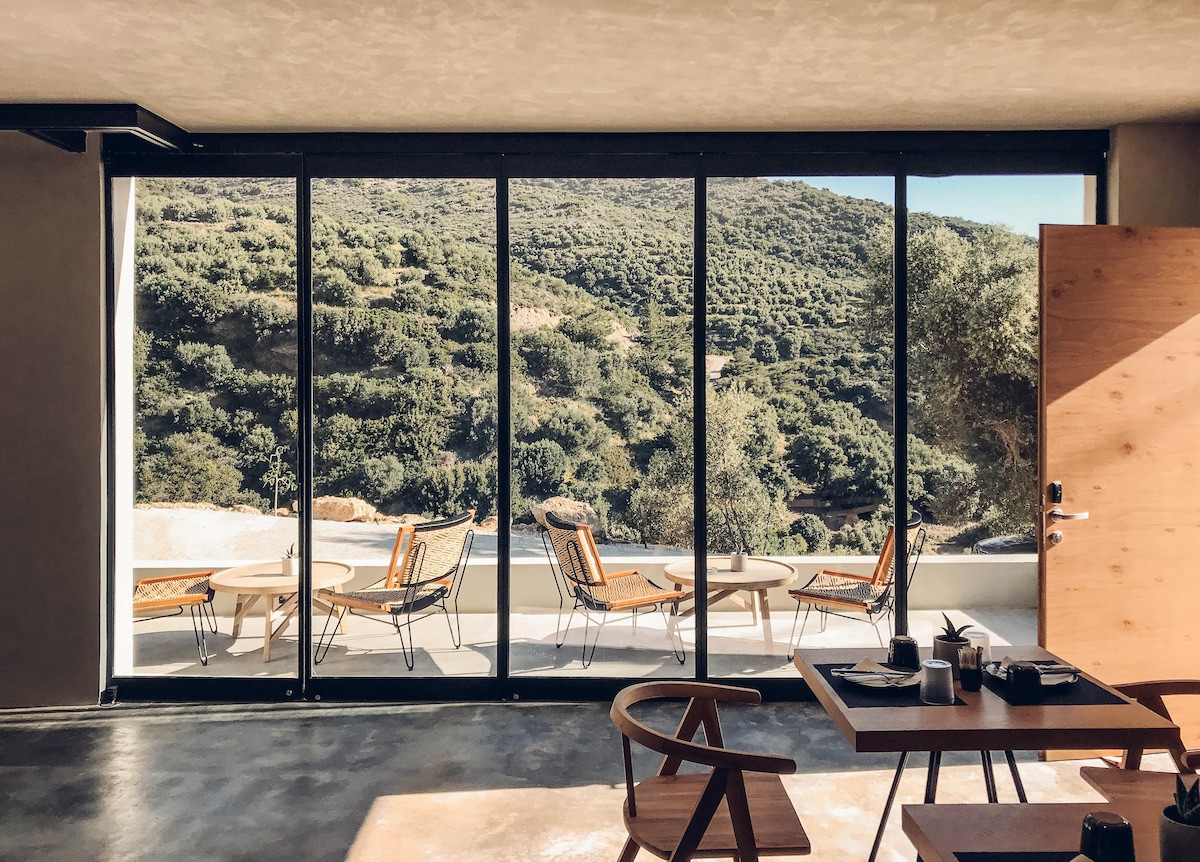 A large glass wall offers a view of the serene landscape beyond, featuring rolling hills covered in greenery. On the spacious balcony, several lounge chairs surround a round table, inviting relaxation. Inside, a wooden dining table with place settings adds a touch of comfort.