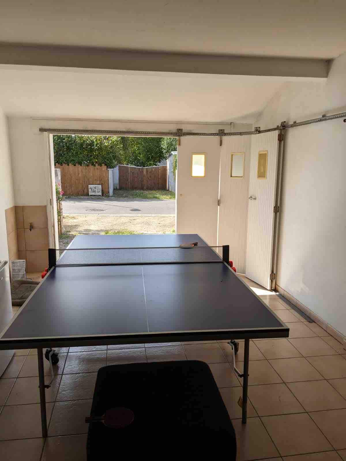 A spacious room is shown with a ping pong table positioned centrally. The walls are painted in a light color, and natural light enters through a doorway, providing visibility to the outdoors. The flooring is tiled, and a black chair is placed nearby.