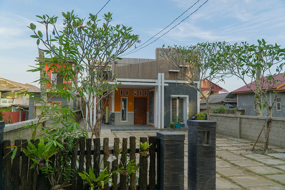 The exterior of the guesthouse features a contemporary design with a combination of gray walls and wooden accents. Lush greenery surrounds the spacious patio area, enhancing the inviting entrance. A stone wall and pathway lead up to the main door, surrounded by well-maintained plants.