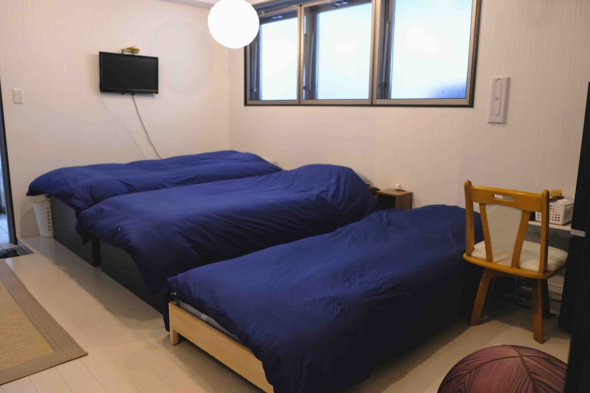 The sleeping area features four spacious tatami beds adorned with navy blue bedding. A wooden chair is positioned near the foot of the beds, while a wall-mounted television is visible above. Natural light filters through several windows, creating a bright atmosphere.