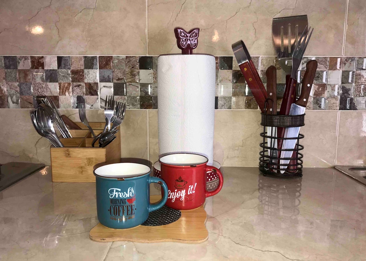 A kitchen countertop displays two ceramic mugs, one blue and one red, situated on a wooden coaster. Nearby, a paper towel holder and a collection of utensils, including forks, knives, and cooking tools, are organized in a metal holder. The surface is clean and well-maintained.
