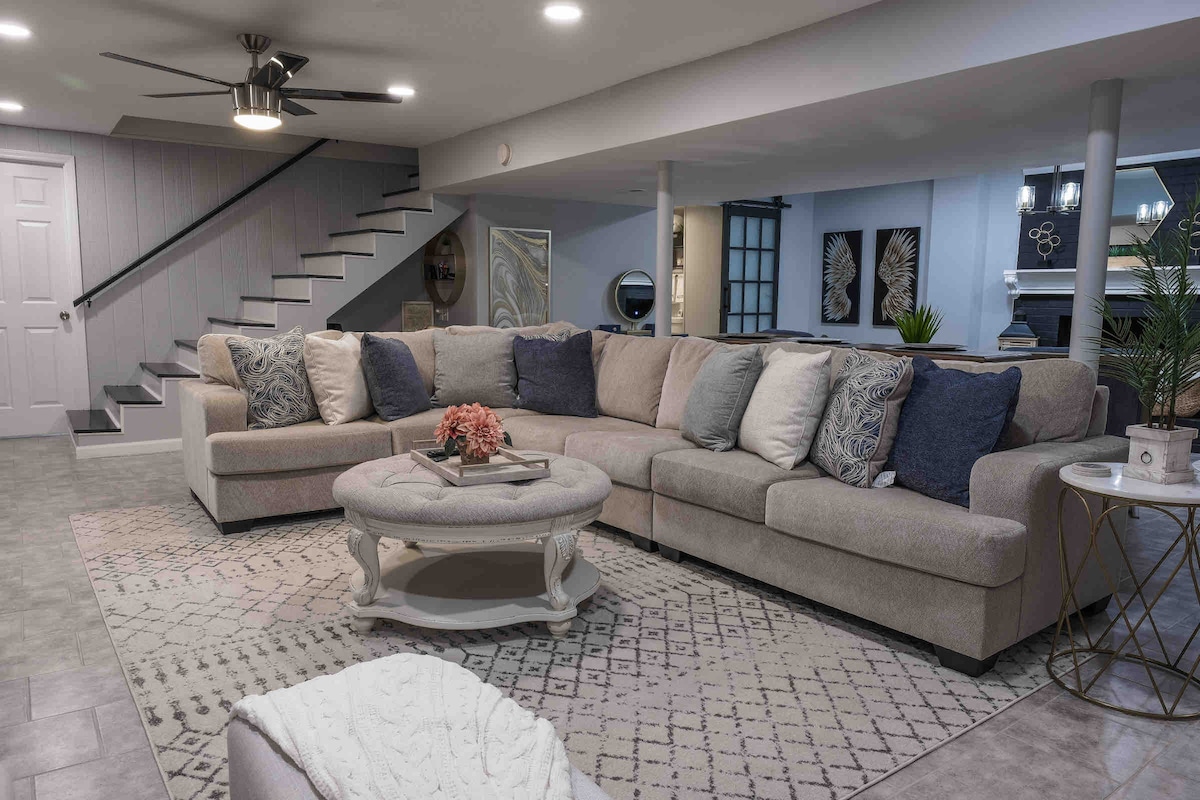 A spacious living room is highlighted by a large sectional sofa, adorned with decorative pillows in various shades of blue and cream. A round coffee table sits in the center, decorated with a floral arrangement. Soft lighting enhances the inviting ambiance of the area.