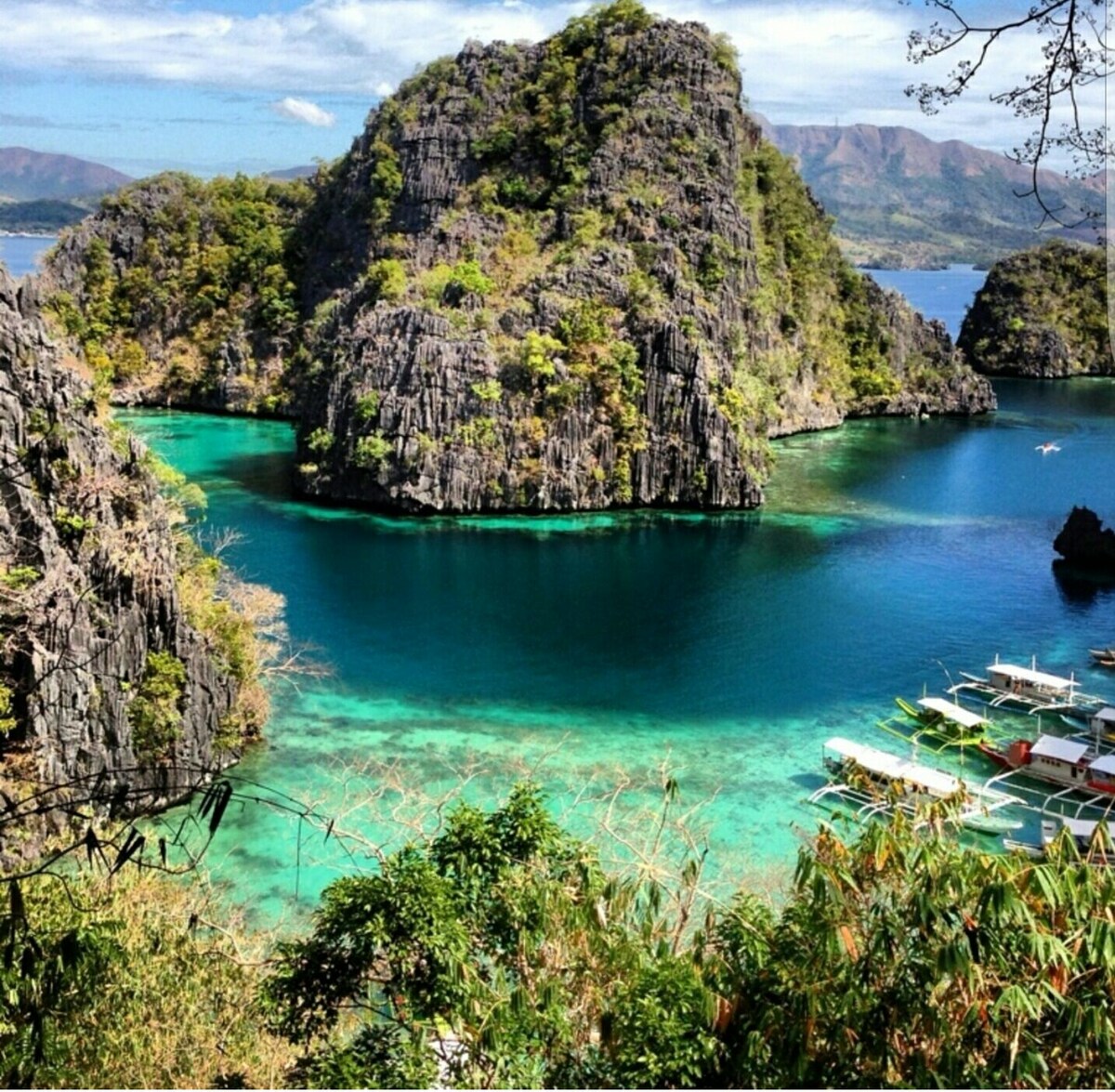 A scenic view of lush, green hills and rocky formations rises above clear turquoise waters. Boats can be seen gently docked along the shoreline, indicating access to nearby tourist destinations. The tranquil landscape is framed by a bright blue sky, enhancing the natural beauty.