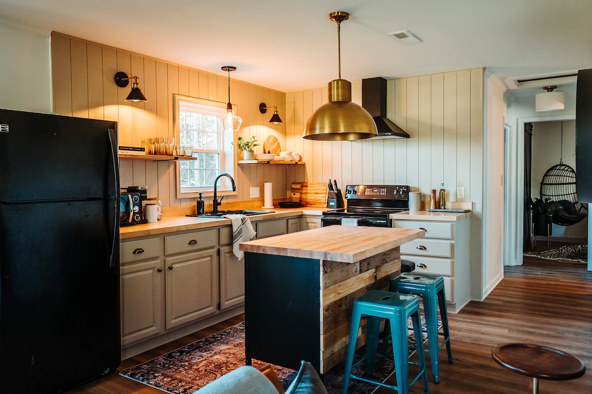 The kitchen features modern cabinetry and a central wooden island with high stools. A large window allows natural light to fill the space, complemented by stylish pendant lighting. Appliances include a black refrigerator, stove, and sink, creating a functional and inviting area for meal preparation.