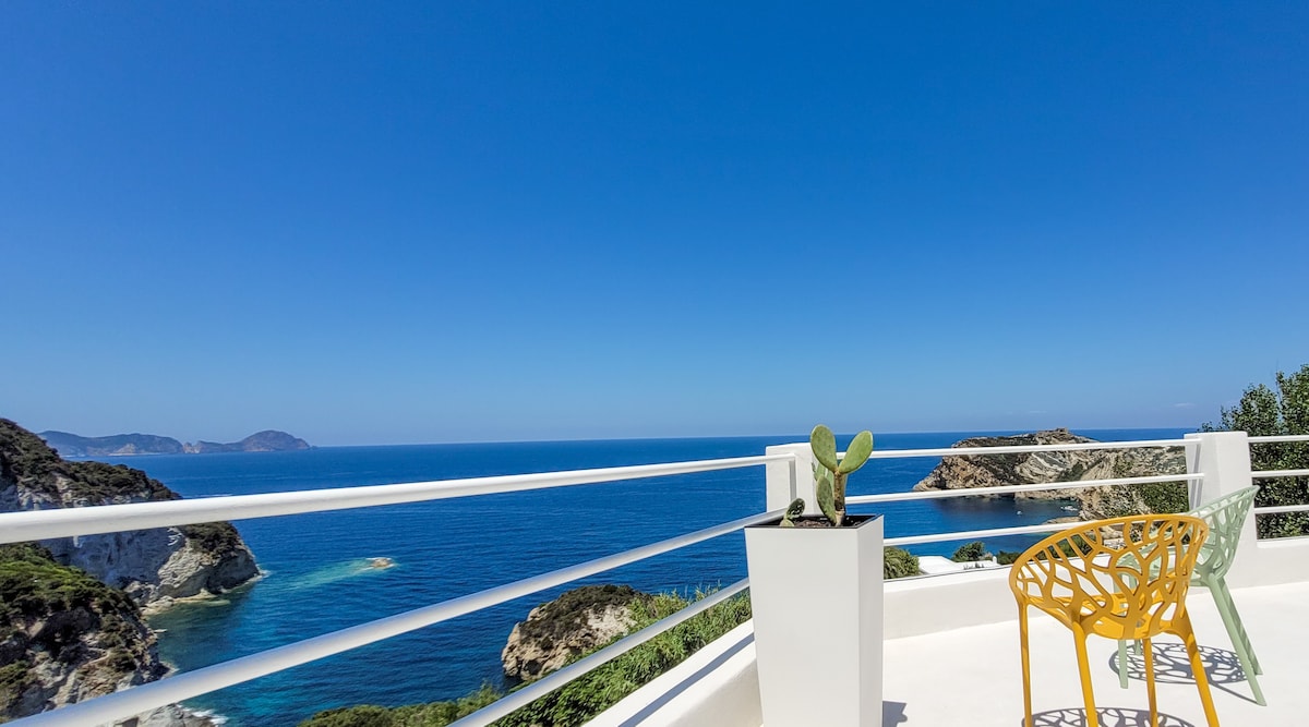 An outdoor terrace offers a sweeping view of the blue sea and distant rocky coastline under a clear sky. A yellow chair and a small potted cactus are positioned on the terrace, enhancing the coastal ambiance.