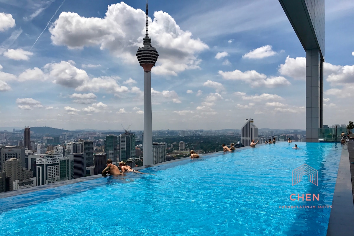 An infinity rooftop pool stretches out against a stunning city skyline, with views of the KL Tower in the background. The clear blue water enhances the serene environment, inviting relaxation. Plush lounge areas are visible along the water's edge, providing space for guests to unwind.