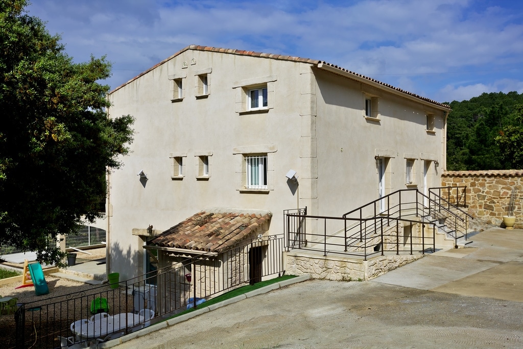 A spacious two-story building is shown, featuring a simple, functional design with white walls and multiple windows. A sloped roof covered in tiles is visible, along with a ramp leading to the entrance. Surrounding areas are paved and well maintained.