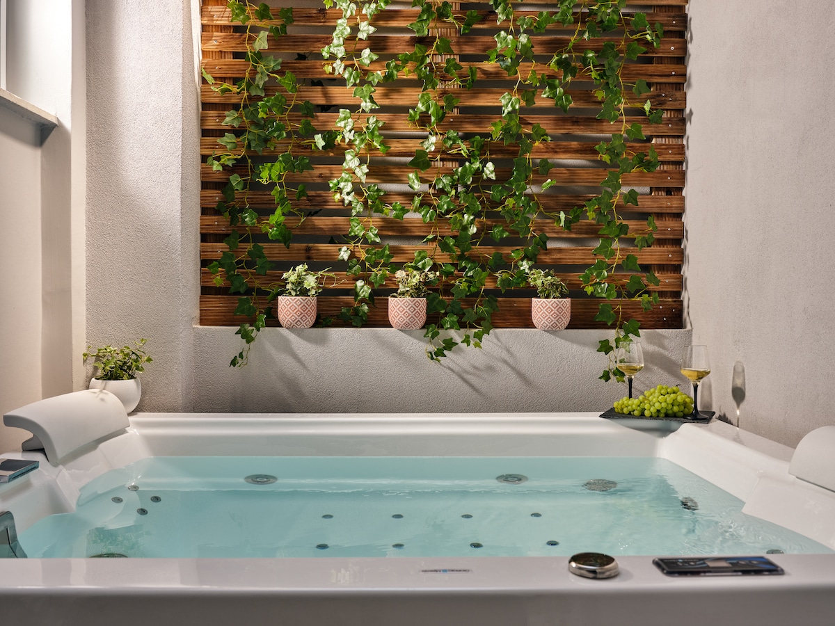 A relaxing hot tub is surrounded by greenery, featuring vibrant plants cascading from a wooden trellis. The water is a soothing blue, complemented by decorative pots and a small tray with wine and grapes nearby.