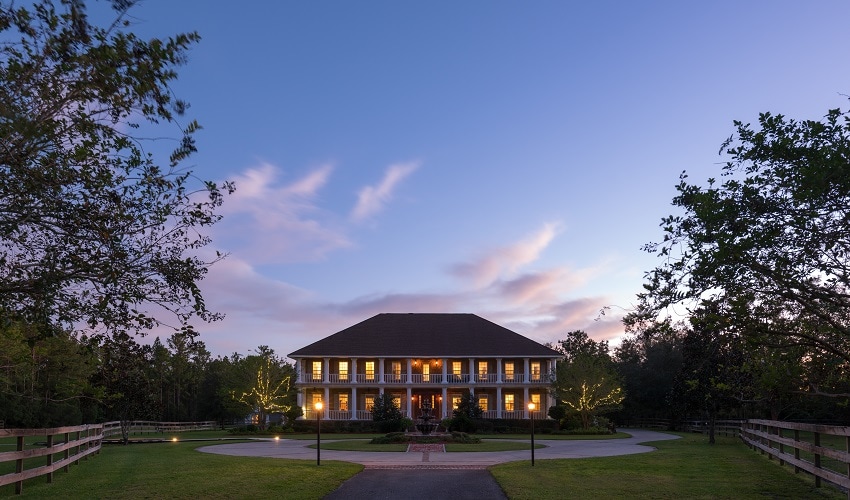 The exterior of the Horse Stamp Inn is displayed at dusk, showcasing a grand two-story structure surrounded by lush greenery. Soft lights illuminate the building and pathways, enhancing the serene atmosphere. A circular driveway allows for easy access, framed by trees and gently lit landscaping.