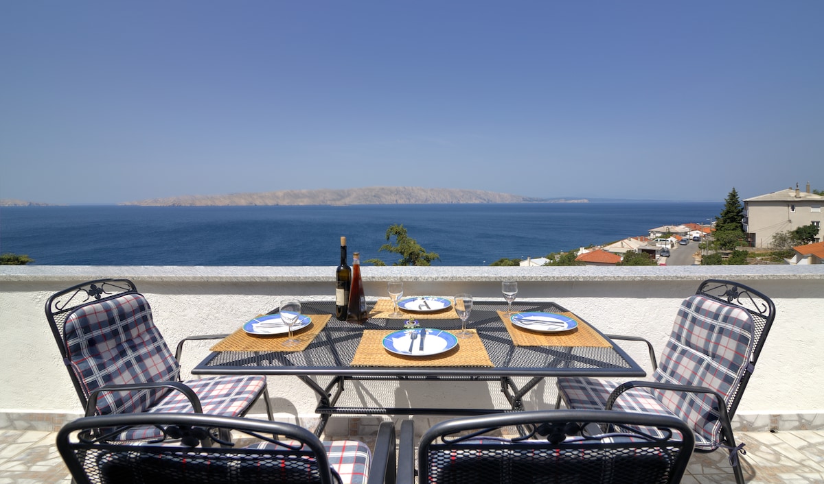 An outdoor dining set is prominently displayed on a balcony, overlooking a panoramic view of the sea and distant coastline. The table is set with plates, glasses, and a bottle, inviting guests to enjoy meals in a scenic environment.