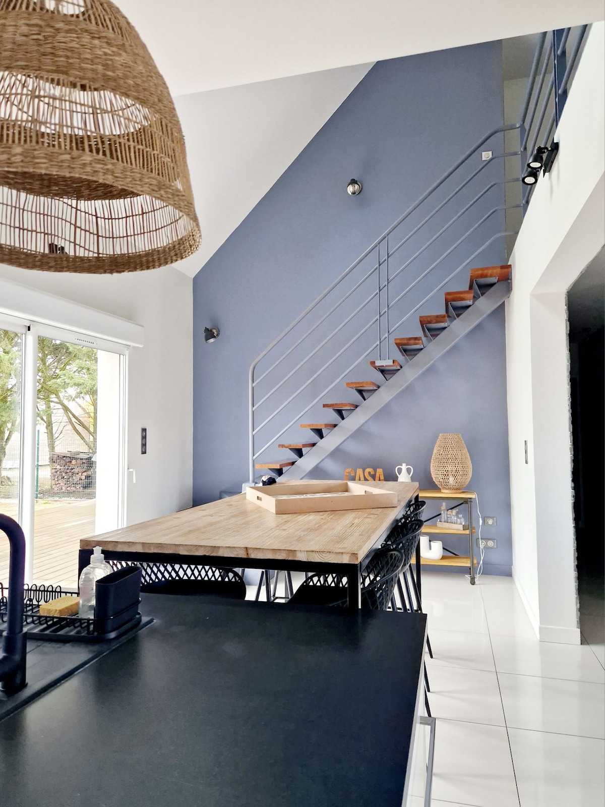An open living area is presented with a large wooden dining table surrounded by black chairs. Light-colored tiles reflect natural light, and a staircase with wooden steps leads to the upper level. The walls are painted in a modern blue tone, enhancing the spacious feel.