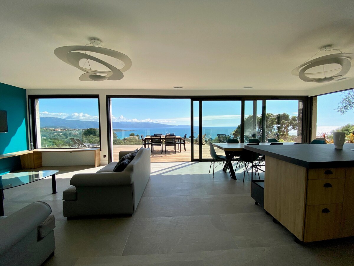 A spacious living area features large glass doors that open to an outdoor dining space, showcasing views of the sea and surrounding hills. Modern furnishings include a dark sofa, a dining table, and a kitchen island that enhance the bright and airy ambiance.