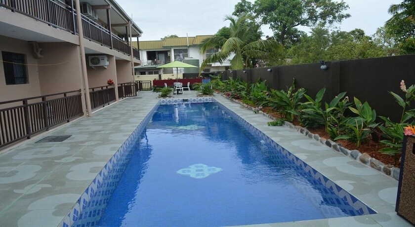 An outdoor swimming pool is surrounded by lush greenery and smooth stone paths. Sun loungers are positioned nearby for relaxation. The building's balconies overlook the pool area, providing a serene and inviting environment.