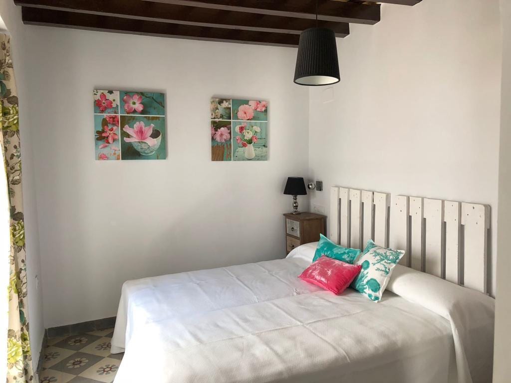 A bright sleeping area features a spacious bed dressed in light bedding. Decorative pillows add a pop of color, while floral artwork adorns the walls. A nightstand with a small lamp complements the room, which is illuminated by natural light filtered through a window with patterned curtains.