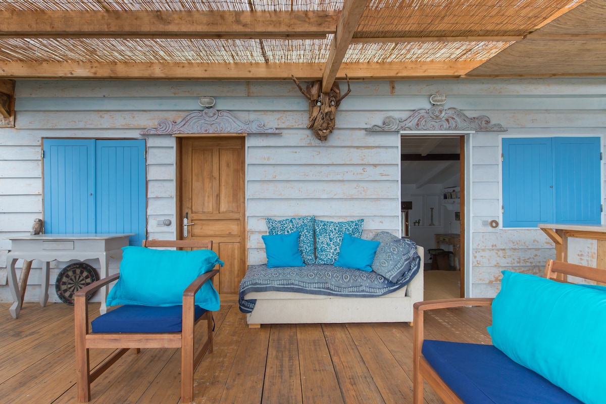 An outdoor terrace features a wooden floor and a rustic ceiling with woven reeds. Blue shutters frame an entrance, while cushions in varying shades of blue adorn a sofa. Several wooden chairs provide additional seating, inviting relaxation in a calm atmosphere.