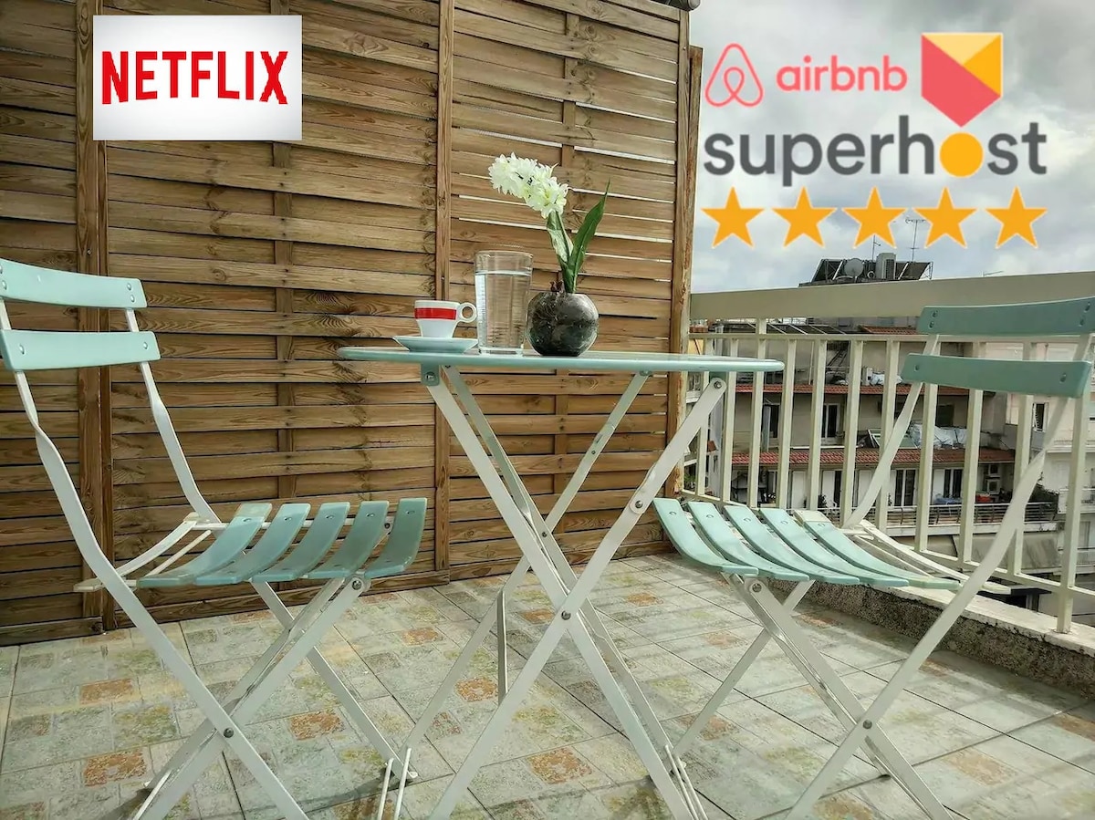 A balcony area features a white metal table paired with two light blue folding chairs. A small vase with white flowers sits on the table, alongside a clear glass of water. The backdrop consists of wooden panels, offering privacy and a cozy atmosphere.