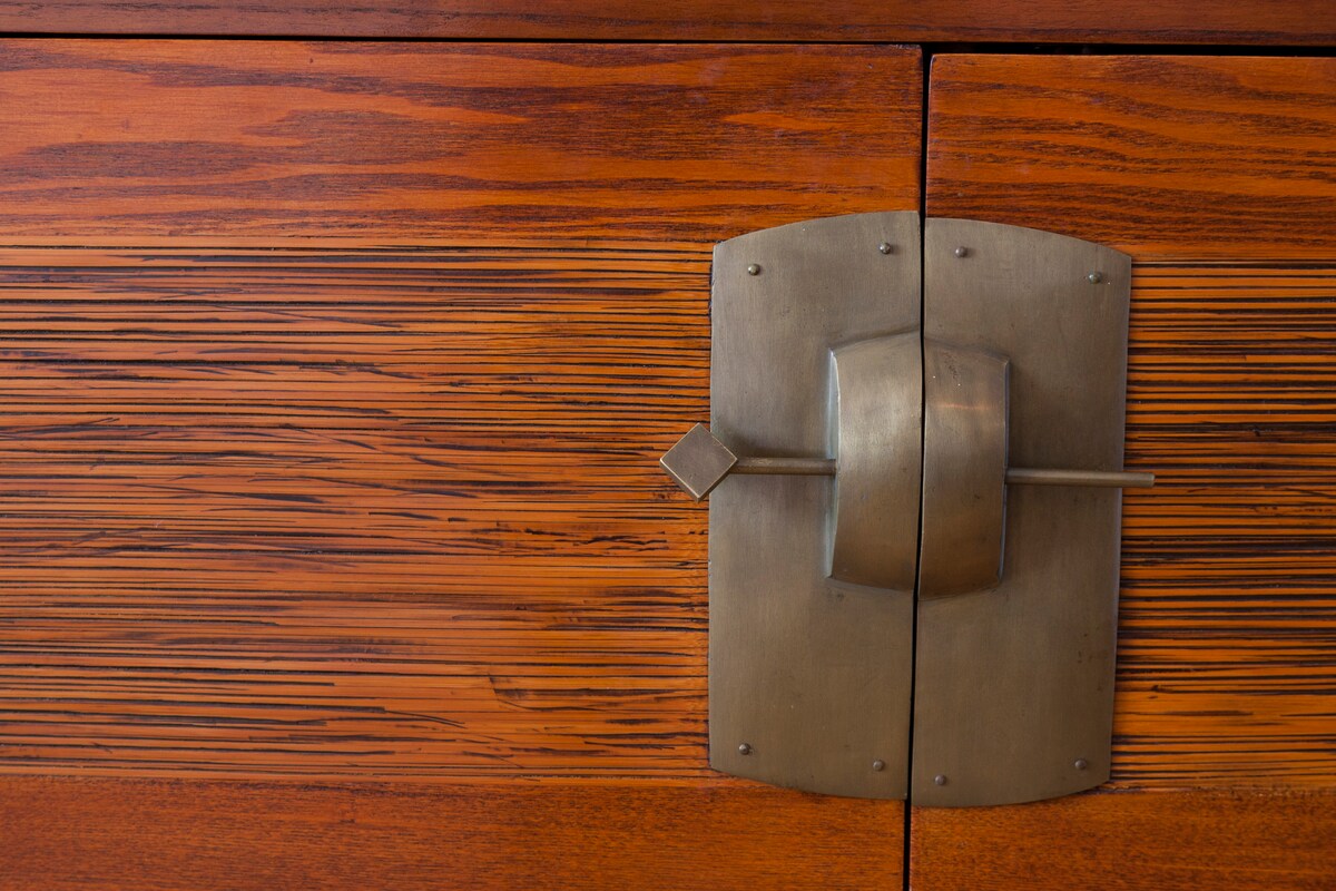 A close-up of a wooden cabinet door showcases a rich, textured grain in warm tones. A rectangular metal latch with a diamond-shaped detail is centered in the middle, contributing to the design's rustic elegance.