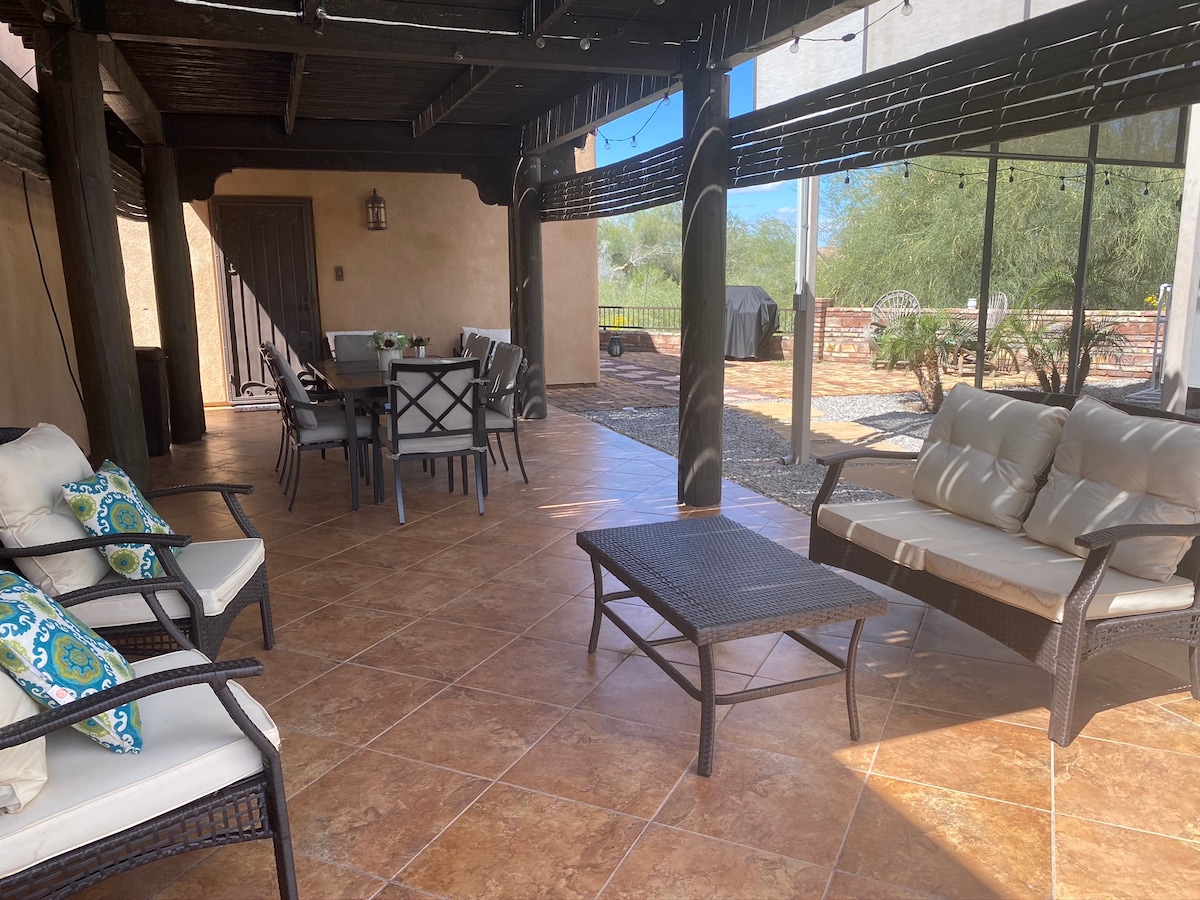 A covered outdoor patio is featured, showcasing a comfortable seating arrangement with plush cushions. A dining table with multiple chairs is positioned nearby. The space boasts tiled flooring and is surrounded by desert landscaping and views of the mountains in the background.