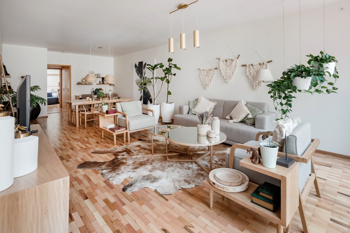 A spacious living room features a combination of neutral-toned furniture and natural wood flooring. Potted plants add greenery to the space, accompanied by cozy seating arrangements. A center table is surrounded by soft rugs, and decorative wall hangings provide a touch of artistry.