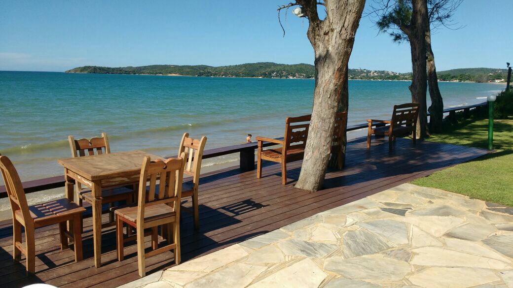A wooden deck facing the beach is highlighted by a dining table surrounded by matching chairs. Nearby, two benches offer additional seating. The calm sea and distant hills create a serene backdrop beneath a clear blue sky.