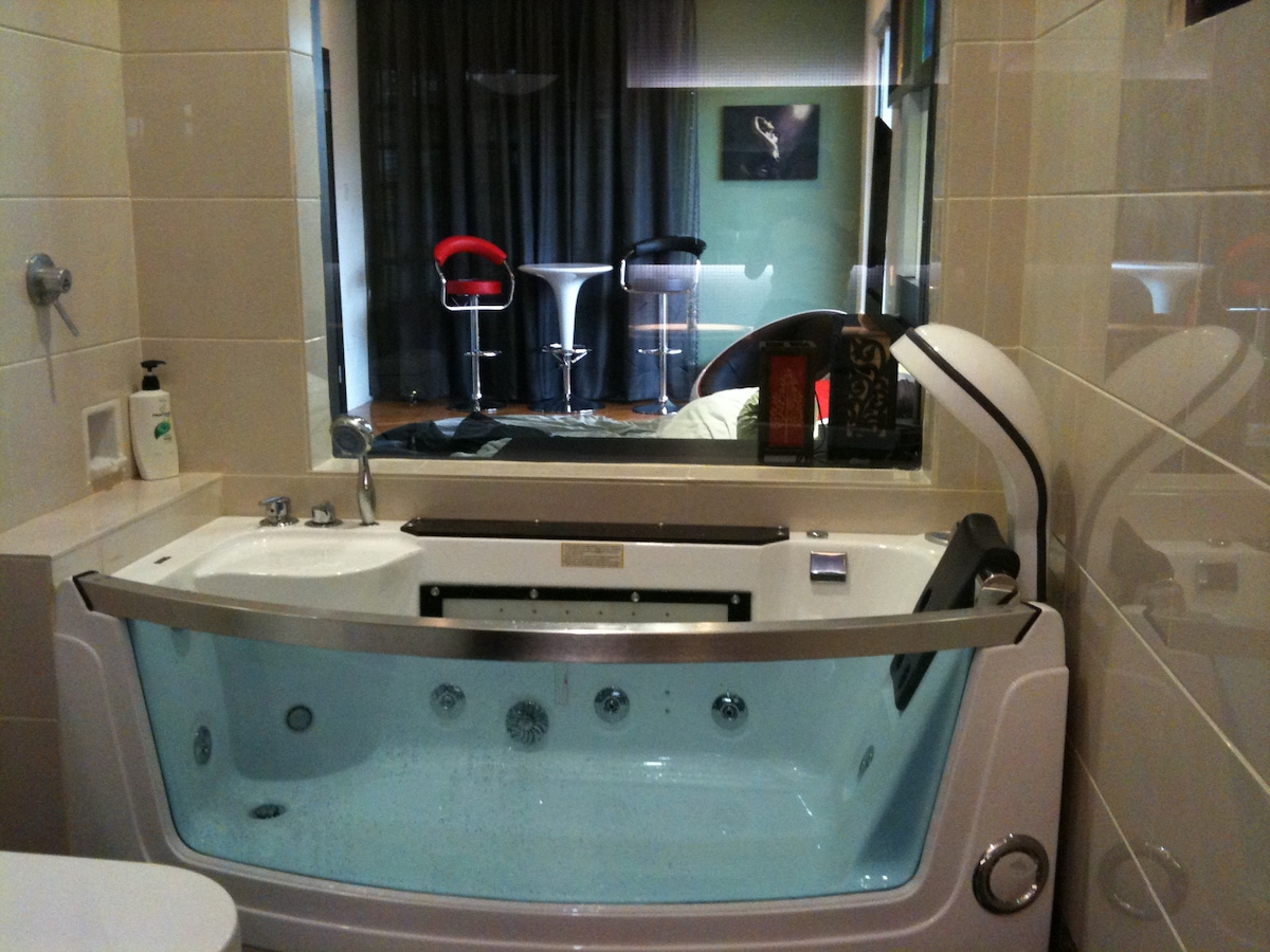 A modern bathroom features a spacious jetted bathtub with a clear view of the adjoining room. A sleek vanity is positioned beside the tub, and stylish bar stools can be seen in the background against a dark curtain. Soft lighting creates an inviting atmosphere.