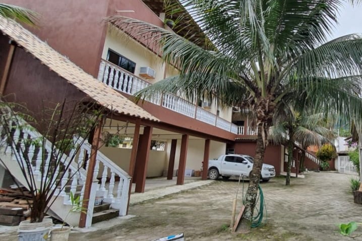 A two-story building is surrounded by palm trees, with a covered parking area visible on the left. Balcony railings are seen on the upper level, while a pathway leads to the entrance underneath. Warm tones of the exterior complement the natural greenery.