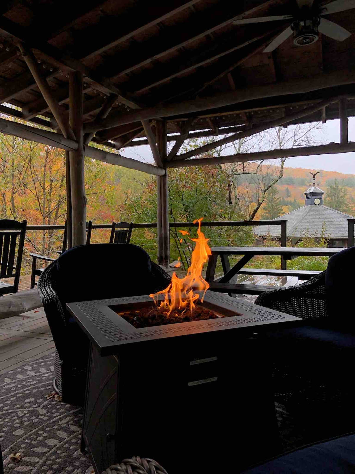 A cozy covered porch features a fire-table with bright flames, surrounded by wooden railings. Comfortable seating is arranged nearby, offering views of the vibrant autumn foliage beyond. The rustic ceiling and natural materials create a warm atmosphere in this serene outdoor space.