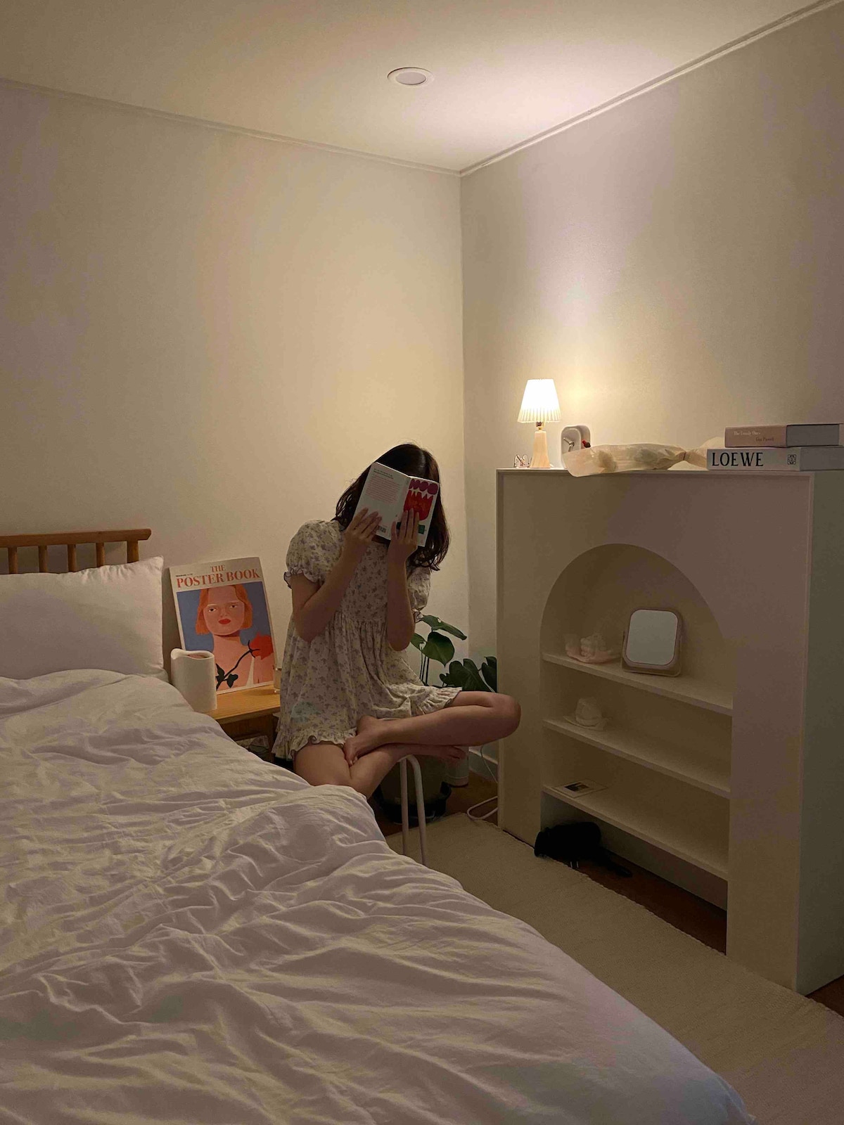 A serene bedroom scene is captured, featuring a spacious bed with a light-colored duvet. A small shelf with books and decorative items is positioned nearby, alongside a lamp casting soft light. A potted plant adds a touch of nature to the cozy atmosphere.