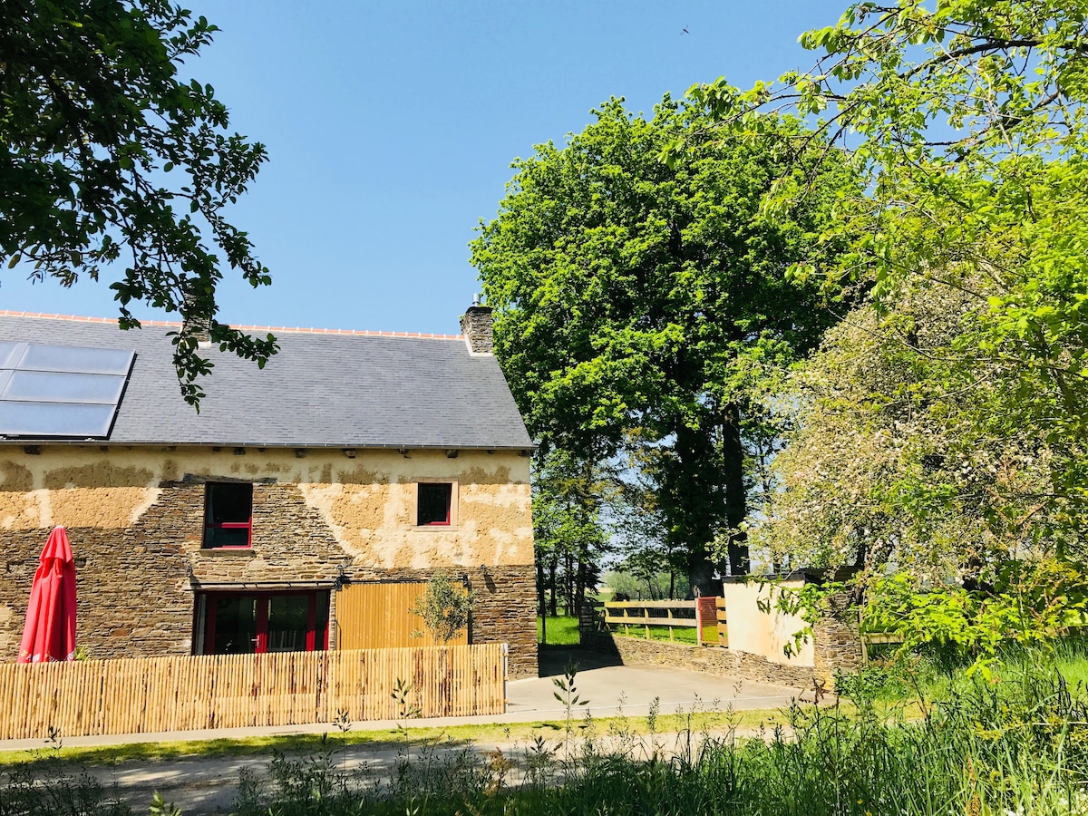 The exterior of the renovated 18th-century longère is shown, featuring a blend of natural stone and rustic wood elements. A vibrant red umbrella provides shade on the terrace, surrounded by lush greenery, with ample blue sky creating a serene atmosphere.