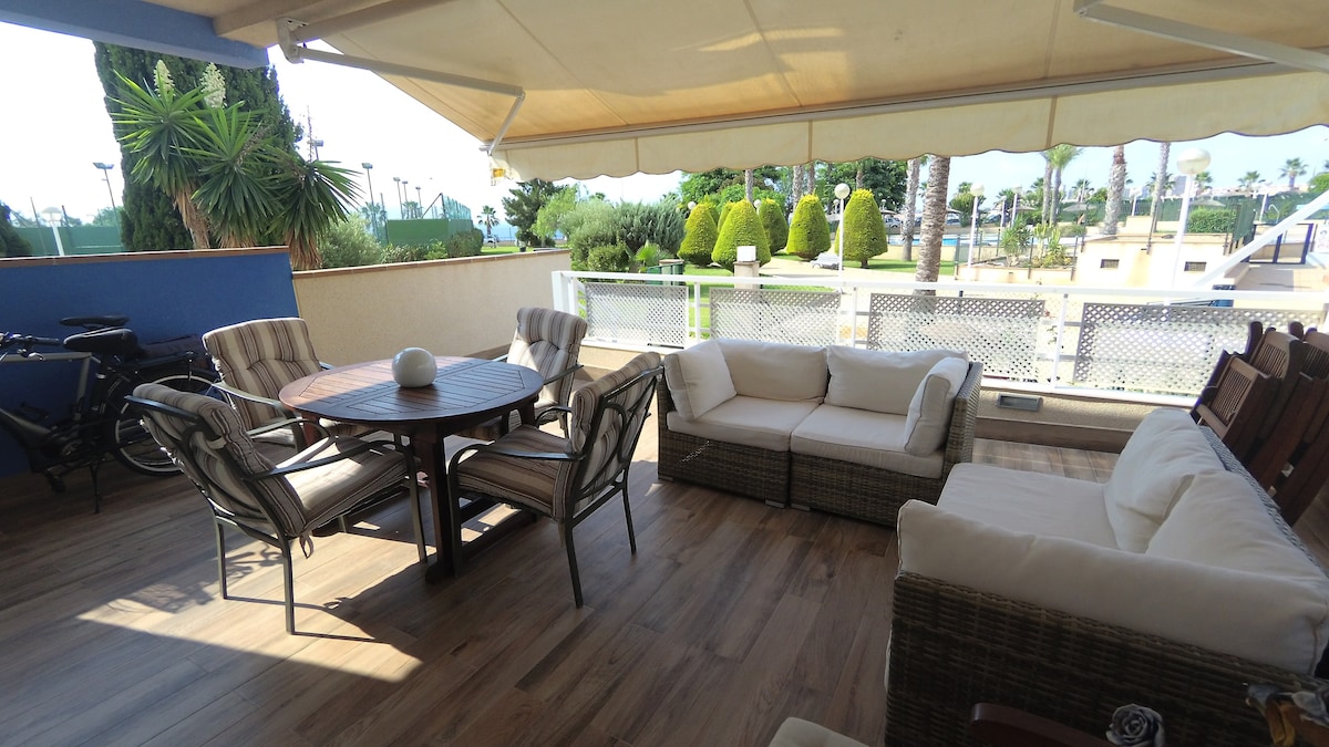 A spacious outdoor balcony area features a round wooden table surrounded by four cushioned chairs. A comfortable sectional sofa is positioned nearby. Potted plants and well-maintained greenery are visible in the background, enhancing the tranquil setting.