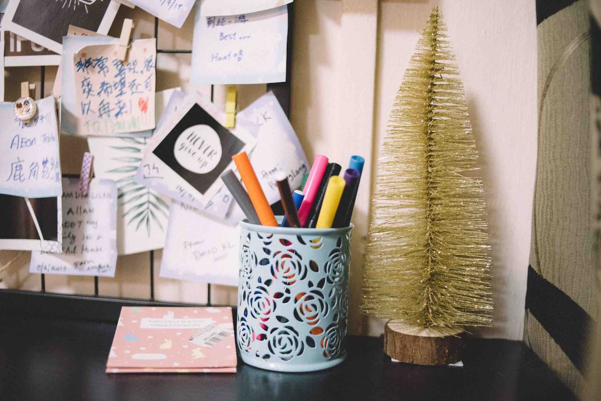 A small decorative arrangement features a golden, glittery Christmas tree alongside a light blue container holding assorted colored markers. Behind them, a notice board is filled with various notes and reminders, adding a personal touch to the workspace.