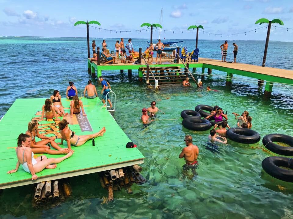 A lively waterfront scene features individuals enjoying leisure time in the clear turquoise water. A vibrant green platform with palm tree decorations serves as a gathering spot, while flotation rings are scattered in the water. A wooden dock is visible, providing access for guests.