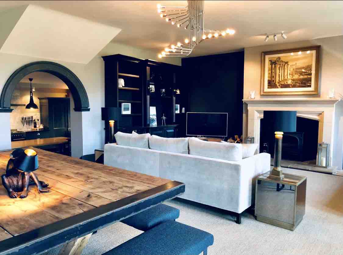 A spacious lounge area features a comfortable sofa with a view towards a stylish fireplace. A large dining table is visible in the foreground, complemented by a striking light fixture overhead. Shelving with decorative items and a television adds to the contemporary decor.