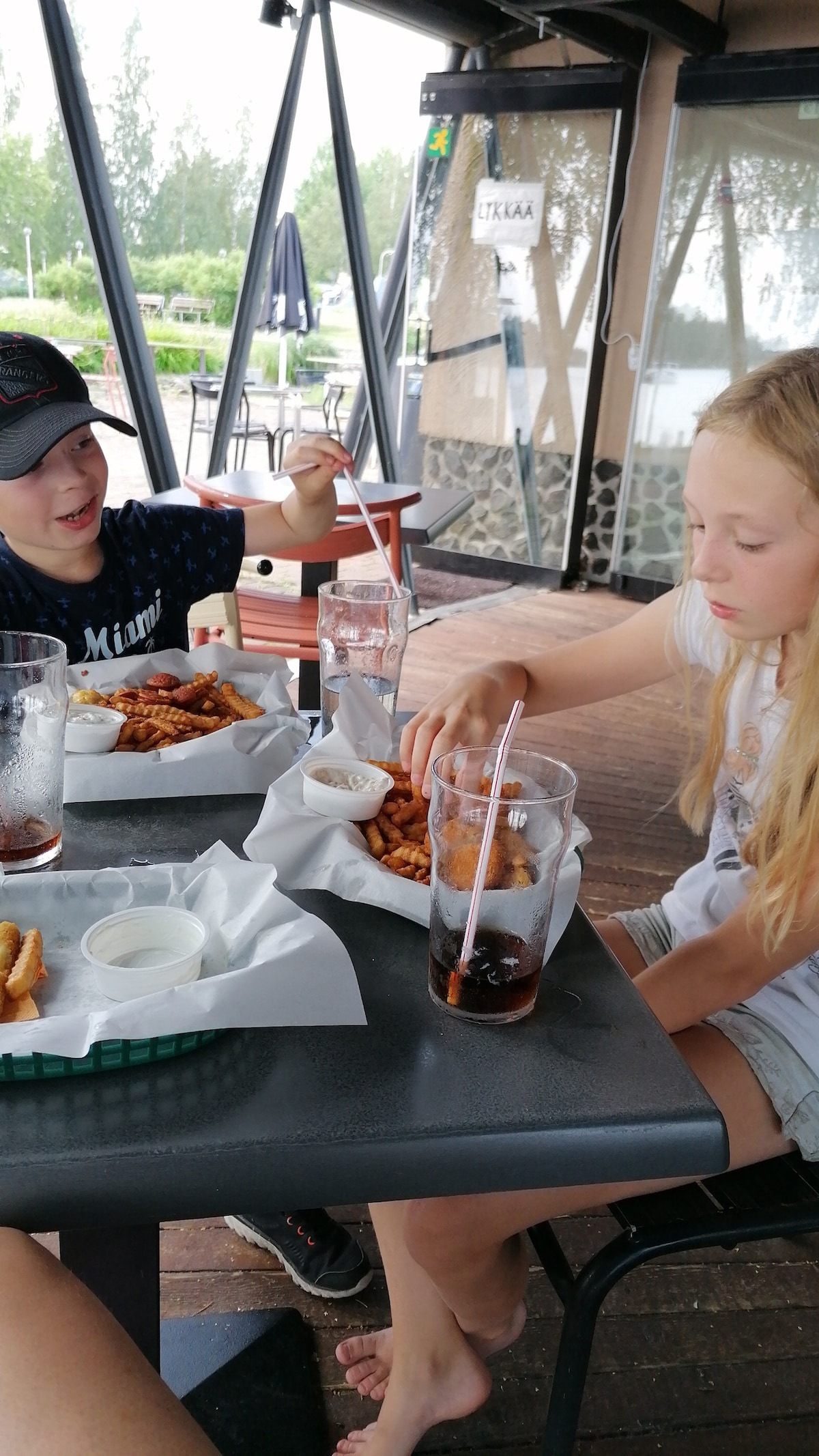A casual dining experience is depicted with two individuals seated at a table. Plates of golden fries are accompanied by drinks in clear glasses. The outdoor seating area is surrounded by large windows, allowing natural light and views of the lush surroundings.