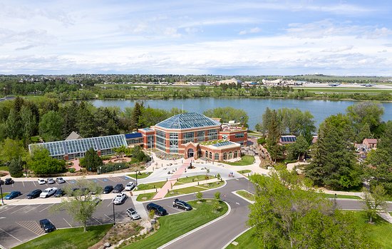 An aerial view captures the vibrant Botanical Gardens, characterized by a large glass conservatory and lush landscaping. A winding path leads through the grounds, bordered by trees and plants, while a serene body of water flows nearby, offering a peaceful setting.