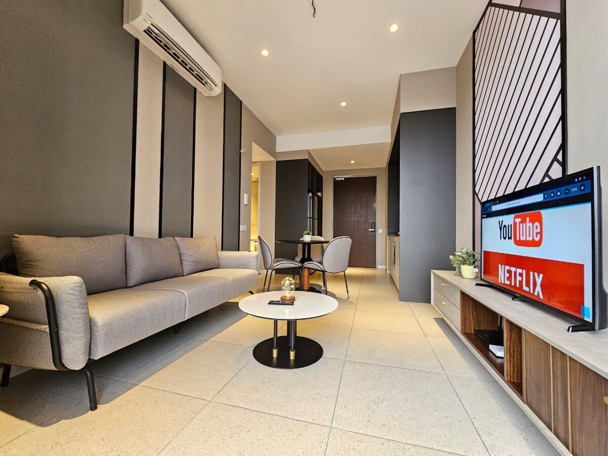 A modern living area features a light gray sofa and a round coffee table on a tiled floor. A flat-screen television is mounted on a wooden console, displaying streaming options. Natural light flows through large windows, enhancing the open and inviting space.