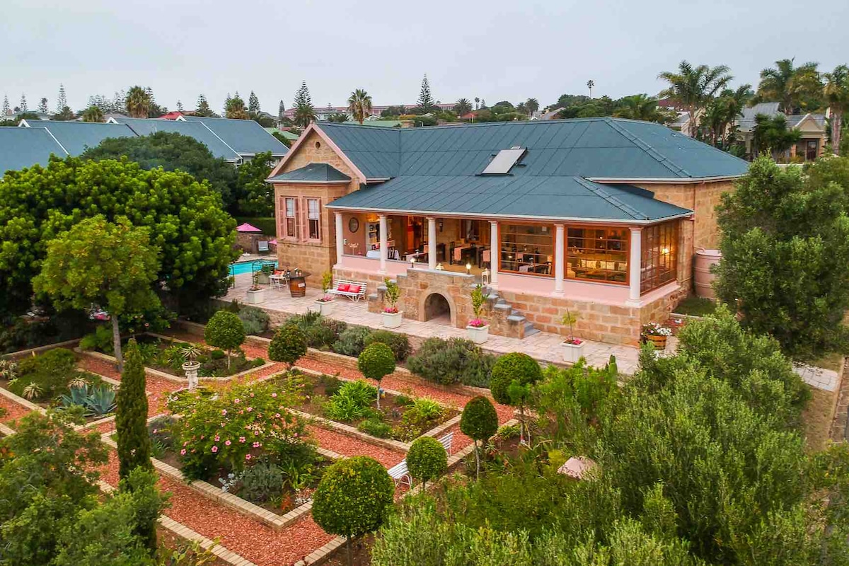 An elegant house with a stone facade and a spacious covered terrace overlooks a beautifully landscaped garden. Neatly trimmed hedges and colorful flowerbeds surround the building. The tranquil pool area is visible in the background, surrounded by lush greenery and palm trees.