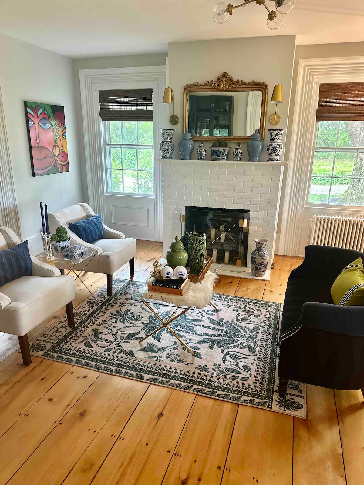 A living room area is shown featuring two light-colored chairs and a dark sofa. A decorative rug sits on the wooden floor beneath a coffee table with greenery. The wall features a mirror and blue vases, while large windows allow natural light to fill the space.
