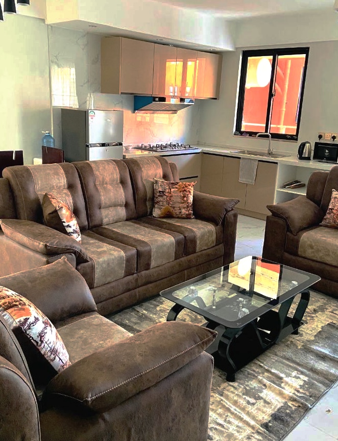 A cozy living area features a brown sectional sofa adorned with patterned cushions, complemented by a glass-topped coffee table. A compact kitchen area is visible in the background, equipped with modern appliances, including a refrigerator and a stove, set against light cabinetry.