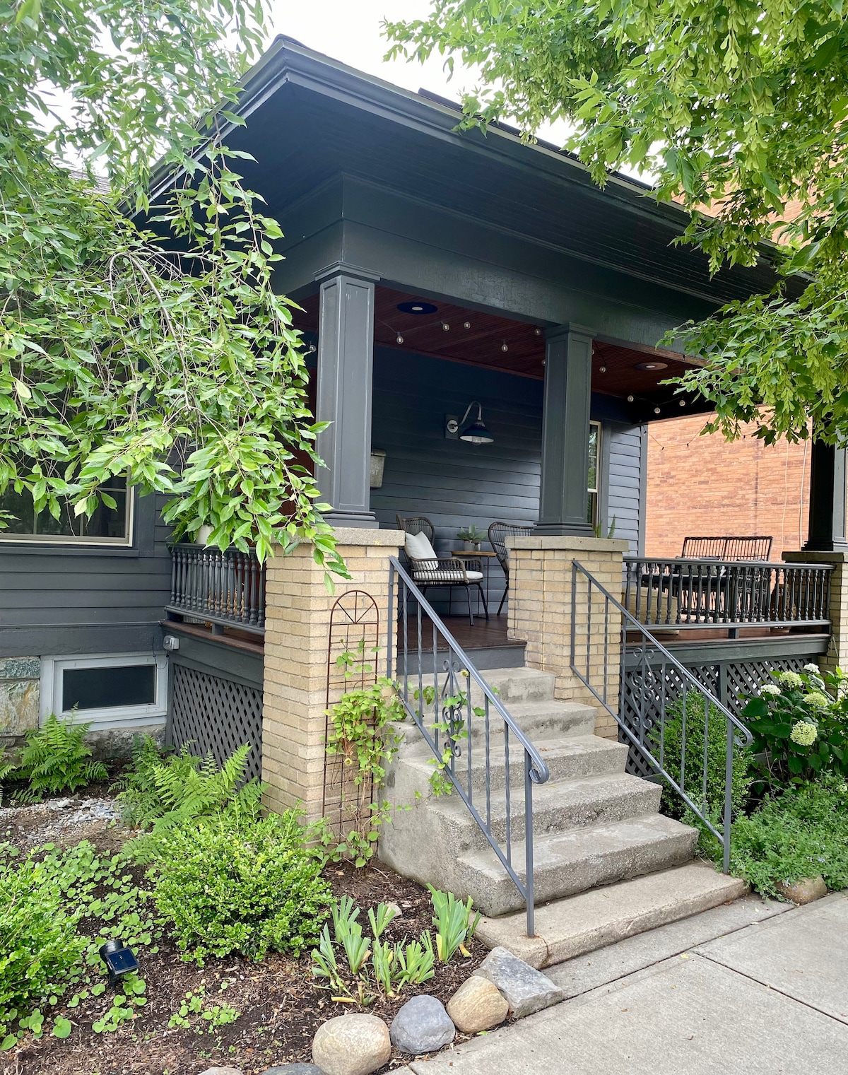 A welcoming front porch is characterized by wood ceilings and comfortable seating arrangements. Stairs lead up to the porch, framed by well-manicured greenery and flowers that enhance the entrance. The structure features a combination of brick and wood siding, contributing to its classic charm.