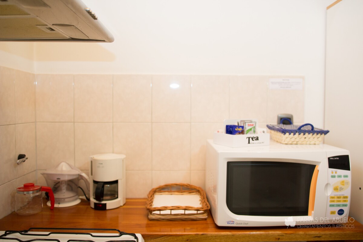 A well-equipped kitchen area features a microwave, a coffee maker, and a compact kettle. Various tea options are neatly arranged in a basket, while a small container holds utensils. Light-colored tiles create a clean backdrop for the wooden counter.