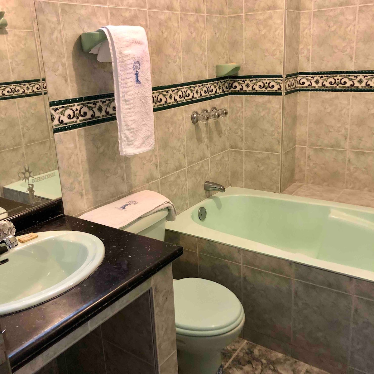 A private bathroom features light green tiles and a matching bathtub, creating a unified look. A black countertop provides a contrasting surface for toiletries. Bath towels are neatly hung on a wall-mounted rack, contributing to the organized space.