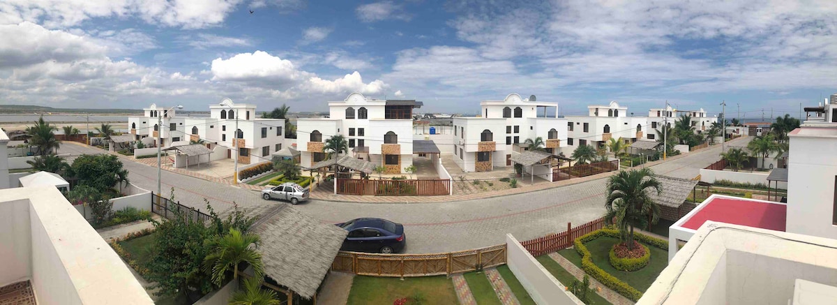 A panoramic view captures the residential community, showcasing white two-story buildings and landscaped gardens. The street is lined with palm trees, and a car is parked along the curb. The scene is framed by greenery in the foreground, enhancing the tranquil setting.