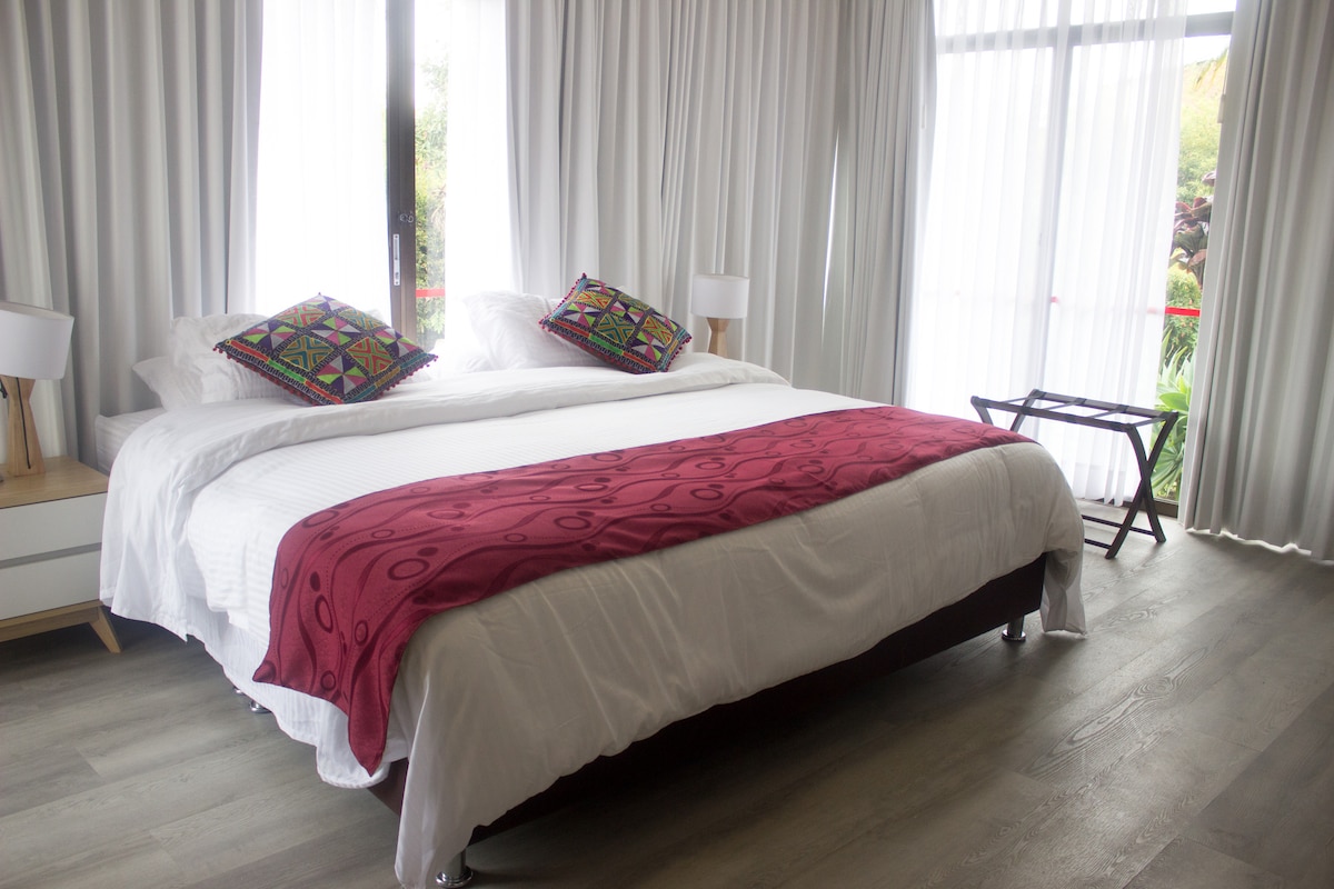 A spacious room features a large bed adorned with a deep red throw. Colorful decorative pillows are placed at the head of the bed. Natural light enters through large glass doors, illuminating the soft white curtains and creating a serene ambiance.
