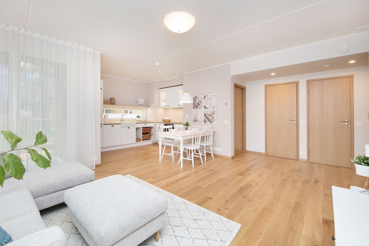A bright and airy living space features a comfortable seating area with a light grey couch. The open kitchen and dining area is equipped with modern fixtures, including a dining table and white chairs. Natural light enters through large windows, illuminating the warm wooden floors.
